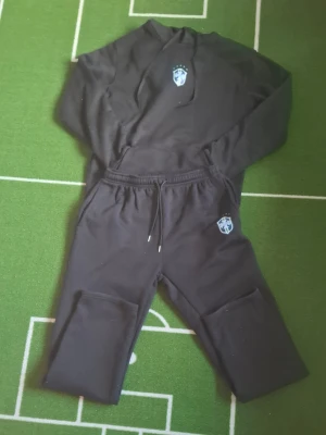 Svart loungewear set med hoodie - Mysigt svart loungewear set med hoodie och matchande byxor. Hoodien har dragsko och en liten blå logga på bröstet. Byxorna har resår i midjan och en ficka på benet. Perfekt för chill hemma eller avslappnade dagar.