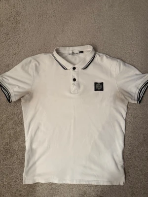 Vit pikétröja från Stone Island - Vit pikétröja från Stone Island med svart logotyp på bröstet. Tröjan har korta ärmar och svarta detaljer på krage och ärmslut. Klassisk pikéknäppning med två knappar och en stilren, sportig look.