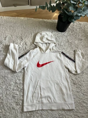 Vit Nike Hoodie - Snygg Nike vit hoodie från Nike med stor röd Swoosh-logga på bröstet och svarta ränder med röda detaljer längs ärmarna. Klassisk känguruficka framtill och huva. Perfekt för en sportig och avslappnad stil. Skriv till mig för fler frågor, svara så snabbt jag kan.🙌