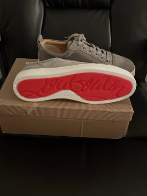 Gråa sneakers med röd sula - Snygga gråa sneakers i mocka från Christian Louboutin med den ikoniska röda sulan och vita kanter. Skorna har klassisk snörning och en stilren design som passar perfekt till streetwear och trendiga outfits.