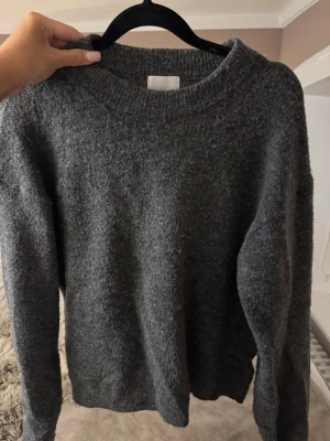 Mörkgrå stickad tröja - Mysig mörkgrå stickad tröja med rund halsringning och långa ärmar. Tröjan har en enkel, klassisk design och är tillverkad i ett mjukt, fluffigt material som känns skönt mot huden. Perfekt att dra på sig när det är kyligt ute.