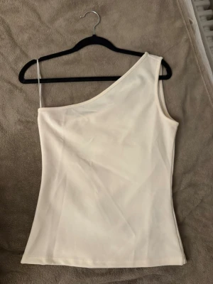 Crémevit oneshoulder topp - Snygg beige topp med oneshoulder-design och smal axelrem på ena sidan. Perfekt för dig som vill ha en stilren och trendig look. Tunn och mjuk i materialet, passar till många olika outfits.