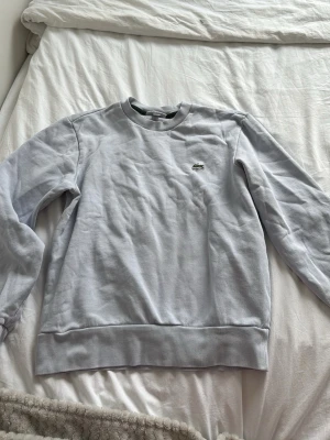 Vit sweatshirt från Lacoste - Klassisk vit sweatshirt från Lacoste med rund halsringning och den ikoniska gröna krokodilloggan broderad på bröstet. Tröjan har långa ärmar och ribbade muddar vid ärmslut och nederkant. Perfekt för en clean och stilren look.