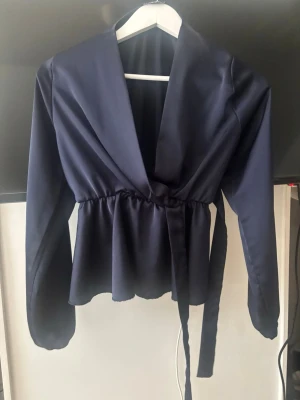 Mörkblå omlottblus med knytband - Snygg mörkblå omlottblus med långa ärmar och knytband i midjan. Blusen har en djup v-ringning och lätt glansigt tyg som ger en elegant känsla. Perfekt att styla med jeans eller kjol för en trendig look.