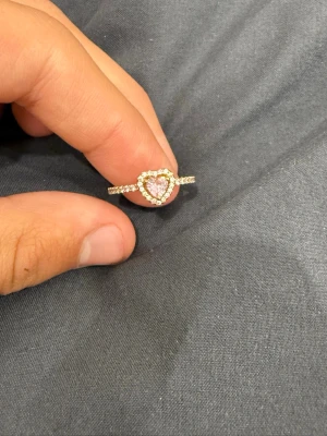 Pandora ring med hjärta och stenar - Säljer en snygg silverfärgad ring från Pandora med ett rosa hjärta i mitten, omgiven av gnistrande vita stenar. Ringen har en tunn band med små stenar längs hela ringen och hjärtat ger en romantisk touch. Perfekt för dig som gillar detaljer och bling.