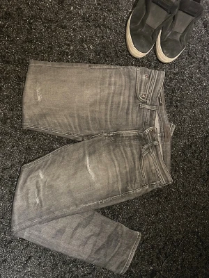 Grå slitna jeans med tvättade detaljer - Säljer ett par grå jeans med slitningar och tvättade detaljer. Jeansen har en smal passform och klassisk femficksdesign. Perfekta för en avslappnad och trendig look. Materialet är denim i bomull.