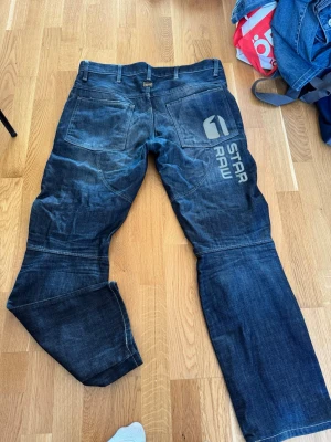 Mörkblå jeans från G-Star Raw 32/34 - Säljer ett par mörkblå jeans från G-Star Raw med stor logga på baksidan av benet. Jeansen har raka ben, stora bakfickor och coola sömdetaljer över knäna. Tillverkade i slitstark denim som ger en snygg streetwear-look.