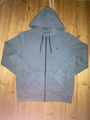 Ralph Lauren grå zip - Snygg grå hoodie med dragkedja och huva. Tröjan har svarta snören i huvan, ribbade muddar och två fickor framtill. Liten broderad logga på bröstet. Perfekt för en avslappnad och stilren look.
