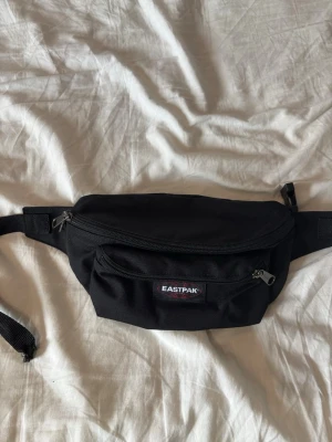 Svart Eastpak midjeväska - Klassisk svart midjeväska från Eastpak med två dragkedjefack och justerbart midjeband. Perfekt för att bära det viktigaste när du är på språng. Väskan har en enkel och stilren design med Eastpak-logga framtill. Aldrig använd