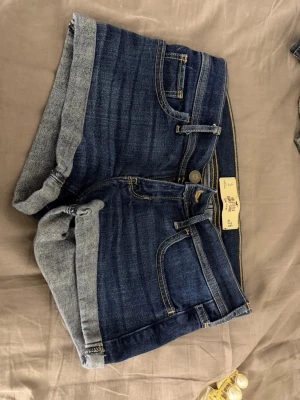 Hollister Mörkblå jeansshorts med uppvik - Snygga mörkblå jeansshorts med uppvikta benslut och kontrastsömmar. Klassisk femficksmodell med gylf och knapp framtill. Perfekta för varma dagar och enkla att matcha med olika toppar.