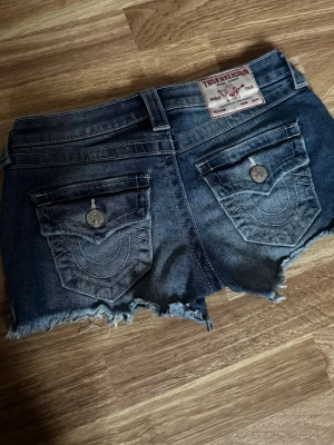Blå jeansshorts från True Religion - Säljer ett par blå jeansshorts från True Religion med lågmidja, fransiga kanter och klassiska bakfickor med lock och dekorativa sömmar. Shortsen har normal passform och bälteshällor. Perfekta för alla tillfällen! Säljer då dom blivit för små men dom är as snygga!! Orginal pris 849kr. Är i super skick har knappt använt dom! Pris går att diskutera💕💕