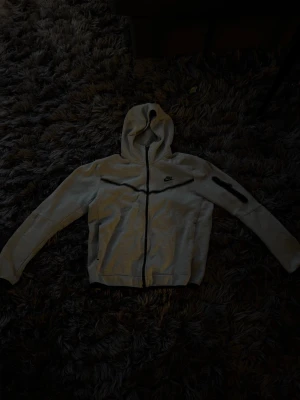 Ljusgrå Nike hoodie med dragkedja - Snygg ljusgrå hoodie från Nike med hel dragkedja framtill och svart Nike-logga på bröstet. Tröjan har huva, långa ärmar och en ficka med dragkedja på ena ärmen. Perfekt för en sportig och avslappnad stil.