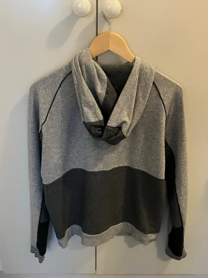 Gråmelerad hoodie med dragkedja - Snygg hoodie i gråmelerat tyg med dragkedja framtill och svart detaljpanel på sidorna. Huva med dragsko och ribbade muddar med randigt mönster vid ärmsluten. Liten BOS-logga på bröstet. Perfekt för en avslappnad och sportig stil.