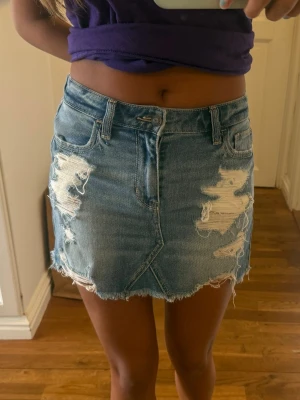 Blå jeanskjol med slitningar - Säljer en kort blå jeanskjol med slitna detaljer och rå kant. Kjolen har klassiska fem fickor och dragkedja fram. Perfekt för dig som gillar en avslappnad och trendig stil.