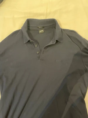 Mörkblå långärmad pikétröja från HUGO BOSS - Snygg mörkblå långärmad polotopp från BOSS med klassisk krage och knappar framtill. Diskret BOSS-logga broderad på bröstet. Perfekt till jeans eller chinos för en clean look.