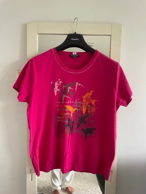 Rosa t-shirt med grafiskt tryck - En snygg rosa t-shirt med korta ärmar och rund hals. Framsidan har ett grafiskt tryck med fåglar i vitt, gult och svart samt textdetaljer. Tillverkad i mjuk bomull som känns skön mot huden. Perfekt för dig som gillar färg och coola prints.