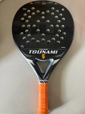 Padelracket Siux Tsunami All Black - Siux, modell Tsunami All Black Limited Edition. Racketen är svart med vita och orange detaljer, har rund form och många hål i träffytan. Handtaget är lindat med orange grepp. Perfekt för dig som vill ta spelet till nästa nivå. Stor sweetspot, skonsamt med bra fart. Nypris 2800