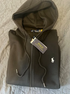 Svart Polo Ralph Lauren kofta - Snygg svart kofta med dragkedja från Polo Ralph Lauren. Klassisk design med huva och dragsnören, samt den ikoniska vita logon broderad på bröstet. Tillverkad i mjukt material som känns skönt mot huden. Perfekt för en avslappnad och stilren look.