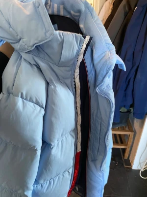 Ljusblå pufferjacka Tommy Hilfiger - Snygg ljusblå pufferjacka från Tommy Hilfiger med hög krage och huva. Jackan har fått ett liten hål vid armen därför har priset sänks även. Jackan har quiltad design, dragkedja framtill och svart innerfoder. Perfekt för kalla vinterdagar och har en modern, sportig look.