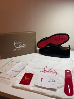 Svarta Louboutin sneakers med nitar - Snygga svarta sneakers från Christian Louboutin med klassisk röd sula och svarta nitar över tån. Skorna har en låg profil, snörning och är tillverkade i mocka. Perfekta för dig som vill sticka ut med en edgy och exklusiv look.