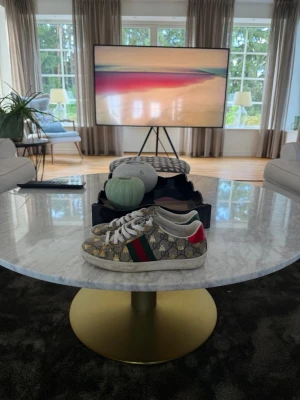 Gucci sneakers med GG-mönster - Snygga sneakers från Gucci med klassiskt GG-mönster i beige och brunt, gröna och röda ränder på sidan samt metallic-detaljer i rött och grönt på hälen. Skorna har vita snören och en låg siluett. Perfekta för dig som vill sticka ut med exklusiv stil.