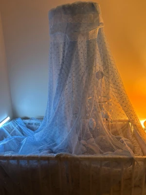 Spjälsäng med blått sänghimmel och dekoration - Säljer en vit spjälsäng med blått sänghimmel i tunn, stjärnmönstrad textil. Inuti finns blått flätat spjälskydd, babykudde, mjuk filt och en mobil med mjuka figurer och träkulor. Dekorativ skylt med texten 'Baby Boy' och söta detaljer ingår.