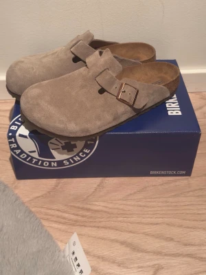 Birkenstock boston - Säljer ett par snygga beige tofflor från Birkenstock i mjuk mocka med justerbart spänne i metall. Klassisk slip-in modell med bekväm fotbädd i kork och mocka. Perfekta för chill dagar hemma eller när du vill ha det extra bekvämt. Pris kan diskuteras!