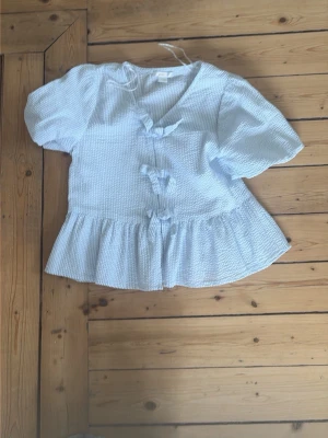 Ljusblå blus med rosetter Gina Tricot - Supersöt ljusblå blus från Gina Tricot med puffiga korta ärmar och tre dekorativa rosetter framtill. Blusen har en v-ringning, volang nedtill och är gjord i ett strukturerat, tunt tyg med diskreta ränder. Perfekt för en trendig och söt look.
