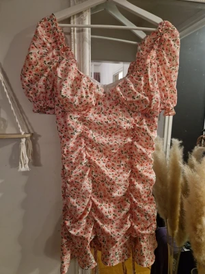 Kort blommig klänning med puffärm - Söt kort klänning med puffärmar och rynkade detaljer. Klänningen är vit med små rosa och gröna blommor, har volangkant nertill och en feminin v-ringning. Perfekt för sommarens alla tillfällen. Storlek M. Passar nog bättre för S eller mindre M.