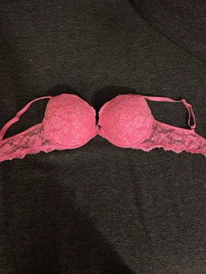 Rosa spets-bh med vadderade kupor - Säljer en snygg rosa bh med vadderade kupor och justerbara axelband. Bh:n är täckt av blommig spets och har en liten rosett mellan kuporna. Materialet är mjukt och stretchigt för skön passform.