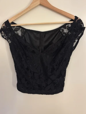 Svart spets topp med v-ringning - Snygg svart topp med v-ringning och spetsdetaljer. Toppen är croppad och har korta ärmar med offshoulder-stil. Perfekt för dig som vill ha en trendig och elegant look. Dragkedja bak för enkel på- och avtagning.