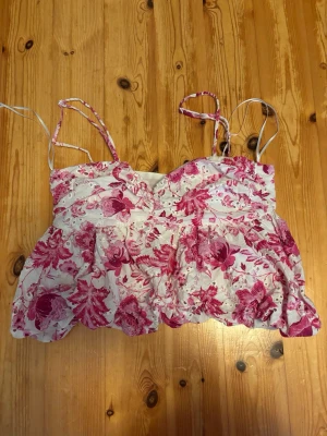 Blommig linnetopp från Zara - Supersöt linnetopp från Zara med rosa blommor på vit botten. Toppen har tunna axelband, volangdetaljer och en luftig passform. Perfekt för varma dagar och ger en romantisk vibe till din outfit.
