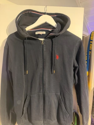 Mörkblå hoodie från U.S. Polo Assn - Mörkblå hoodie med dragkedja från U.S. Polo Assn. Klassisk design med huva och dragsnören, två fickor framtill och röd logga broderad på bröstet. Insidan av huvan har en snygg randig detalj i rött, vitt och blått. Perfekt för en chill och sportig look.