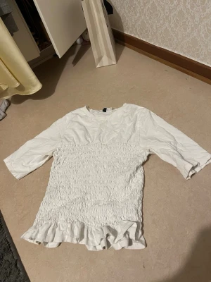 Vit topp med volang från H&M Divided - Supersöt vit topp från H&M Divided med smockad midja och volang nertill. Toppen har trekvartsärm och är gjord i mjuk bomull. Perfekt för en trendig och avslappnad look.