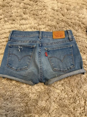 Blå jeansshorts från Levi's - Säljer ett par klassiska blå jeansshorts från Levi's med uppvikta benslut och två bakfickor, varav ena har en liten slitning. Shortsens ikoniska läderpatch och röda Levi's-tag syns tydligt. Perfekta för sommaren! Gjort om själv från jenas till shorts