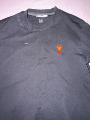 Svart t-shirt från Ami Paris - Svart t-shirt från Ami Paris med klassisk passform och rund hals. På bröstet finns Ami:s ikoniska röda hjärta med A-broderi. Perfekt basplagg för dig som gillar stilrena detaljer och hög kvalitet.