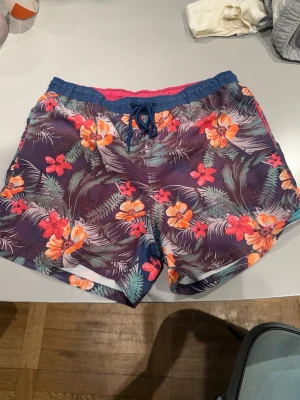 Blommiga badbyxor med dragsko - Säljer ett par färgglada badbyxor med tropiskt blommönster i orange, rött och grönt på mörkblå botten. De har elastisk midja med blå dragsko och en bakficka. Perfekta för stranden eller poolen!