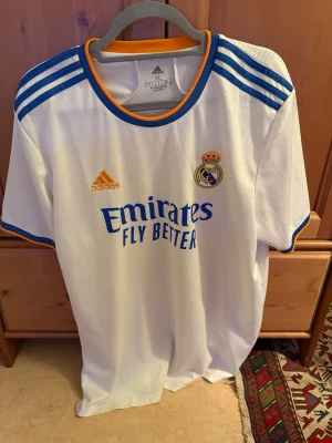 Real Madrid matchtröja Adidas XL - Säljer en vit Real Madrid matchtröja från Adidas i storlek XL. Tröjan har blå detaljer på axlarna, orange kant vid hals och ärmslut samt klubbmärke och Adidas-logga på bröstet. Tillverkad i lätt och ventilerande material, perfekt för fotboll eller träning.