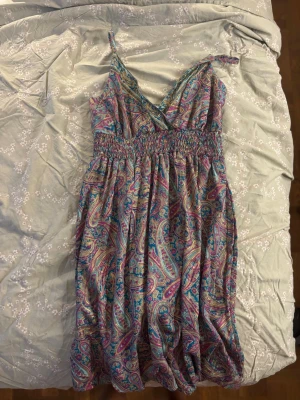 Färgglad paisleymönstrad klänning - Säljer en färgglad klänning med paisleymönster i blått, rosa och gult. Klänningen är tillverkad i 70% konstsilke och 30% polyester, vilket ger ett mjukt och glansigt intryck. Perfekt för dig med lite större byst, den var lite för stor för min.