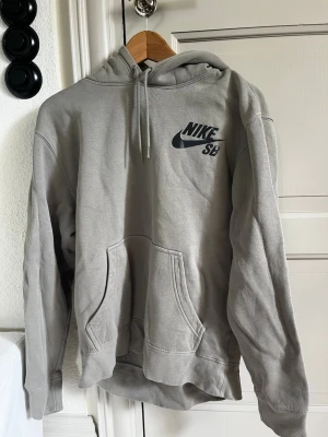 Grå Nike SB hoodie med huva - Säljer en grå hoodie från Nike SB med klassisk logga på bröstet. Hoodien har huva med dragsko och en stor magficka. Tillverkad i mjukt material som passar perfekt för en avslappnad stil.