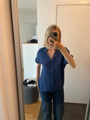 Marinblå kortärmad blus med v-ringning - Marinblå blus med korta ärmar och djup v-ringning. Blusen har en avslappnad passform och är tillverkad i ett lätt, luftigt tyg. Aldrig använd. 15 kr!