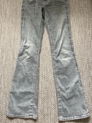 Grå bootcut jeans med slitningar - Ett par grå jeans med bootcut-modell och snygga slitningar längs benen. Jeansen har klassisk femficksdesign och är tillverkade i denim. Perfekta för en avslappnad och trendig look.