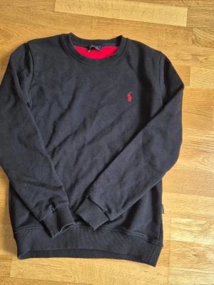 Svart sweatshirt  - Klassisk svart sweatshirt  med röd broderad logga på bröstet. Tröjan har rund halsringning, ribbade muddar och är tillverkad i mjuk bomull. Perfekt för en avslappnad och stilren look.