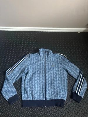 Blå mönstrad ziptröja Lacoste - Säljer en blå ziptröja från Lacoste med geometriskt mönster och vita ränder längs ärmarna. Tröjan har hög krage, muddar i mörkblått och dragkedja framtill. Klassisk krokodillogga på bröstet.