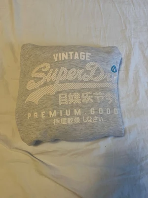 Grå Superdry hoodie med tryck - Snygg ljusgrå hoodie från Superdry med stort vitt tryck på bröstet.  Perfekt för en avslappnad och trendig stil. Tillverkad i mjukt material för extra komfort.