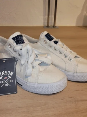 Vita låga sneakers från Vinson Polo Club - Snygga vita sneakers från Vinson Polo Club med klassisk låg profil. Skorna har vita snören, silverfärgade öljetter och en logga på sidan samt plösen. Tillverkade i canvasmaterial med vit gummisula och diskreta detaljer.