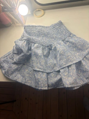 Blommig volangkjol i blått - Supersöt kort kjol med volanger i ljusblått tyg och små vita blommor. Kjolen har bred smock i midjan för skön passform och är perfekt för vår och sommar. Tillverkad i ett lätt och luftigt material som känns bekvämt hela dagen.