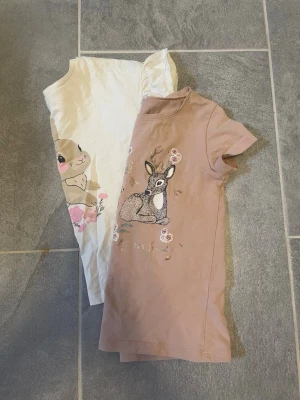 Två söta t-shirts med djurmotiv för barn - Två t-shirts för barn i mjuk bomull, en i beige med tryck av rådjur och blommor, och en i vit med kanin och rosa blommor. Båda har korta ärmar och rund hals. Perfekta för dig som gillar gulliga motiv och vill ha något bekvämt och stilrent.