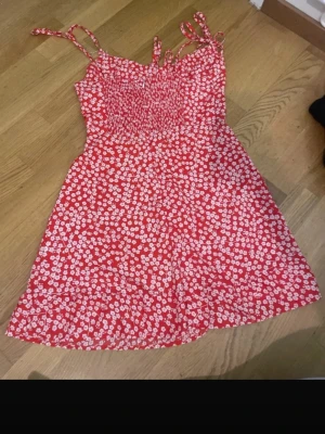 Röd blommig kortklänning med knytband - Söt röd kortklänning med vita blommor och smockad överdel. Klänningen har tunna axelband med knyt och en lätt utställd kjol. Perfekt för varma dagar och ger en somrig vibe.