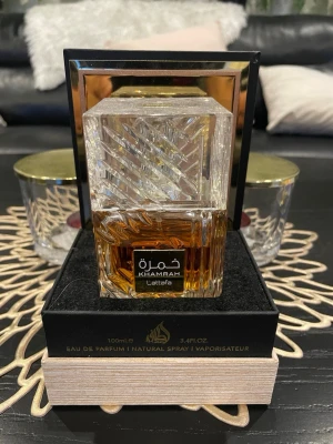 Lattafa Khamrah Eau de Parfum 100ml - Lattafa Khamrah Eau de Parfum i en lyxig, tung glasflaska med geometriskt mönster och guldiga detaljer. Flaskan har en fyrkantig form och innehåller 100 ml av en bärnstensfärgad parfym. Perfekt för dig som gillar exklusiva och orientaliska dofter.